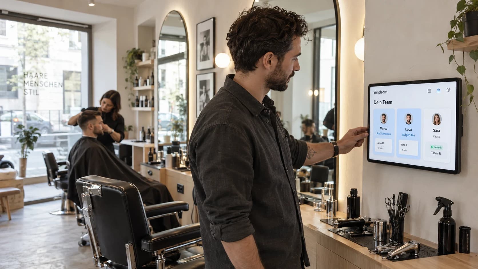 Ein Salonmitarbeiter bedient das SimpleCut-Dashboard auf einem Wand-Tablet, während im Hintergrund ein Kunde einen Haarschnitt erhält.